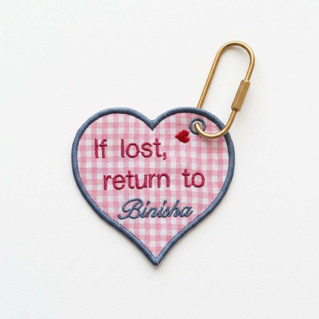 Personalized 'If lost, return to Binisha' embroidered heart bag tag on pink gingham with blue border.