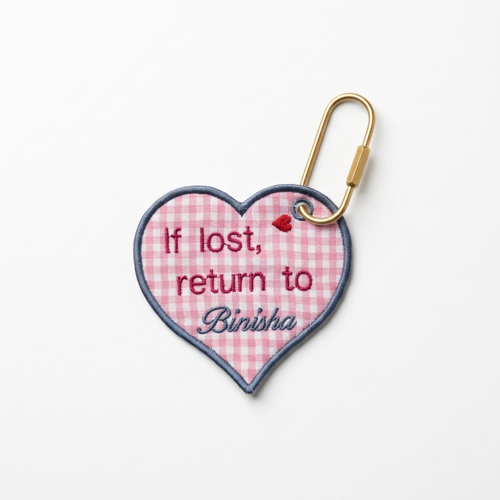Personalized 'If lost, return to Binisha' embroidered heart bag tag on pink gingham with blue border.